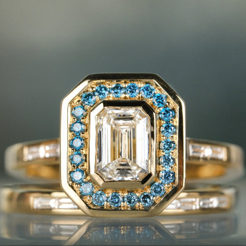 Gold ring with emerald-cut center stone and blue gemstone accents on a reflective surface.
