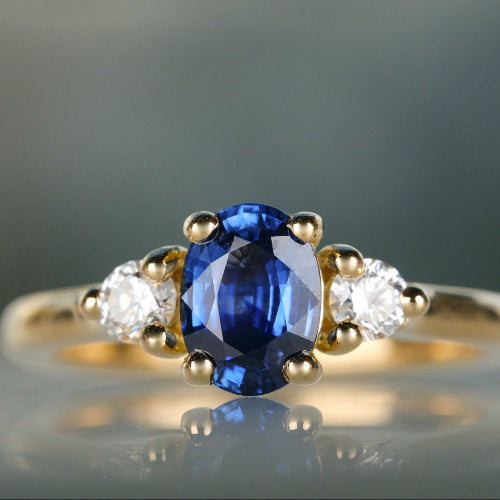 Gold ring with a blue gemstone and two white stones on a reflective surface.