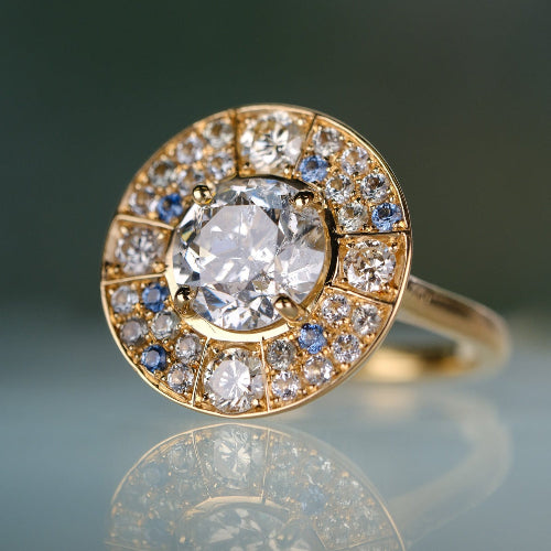 Gold ring with a large central diamond and smaller diamonds around on a reflective surface.