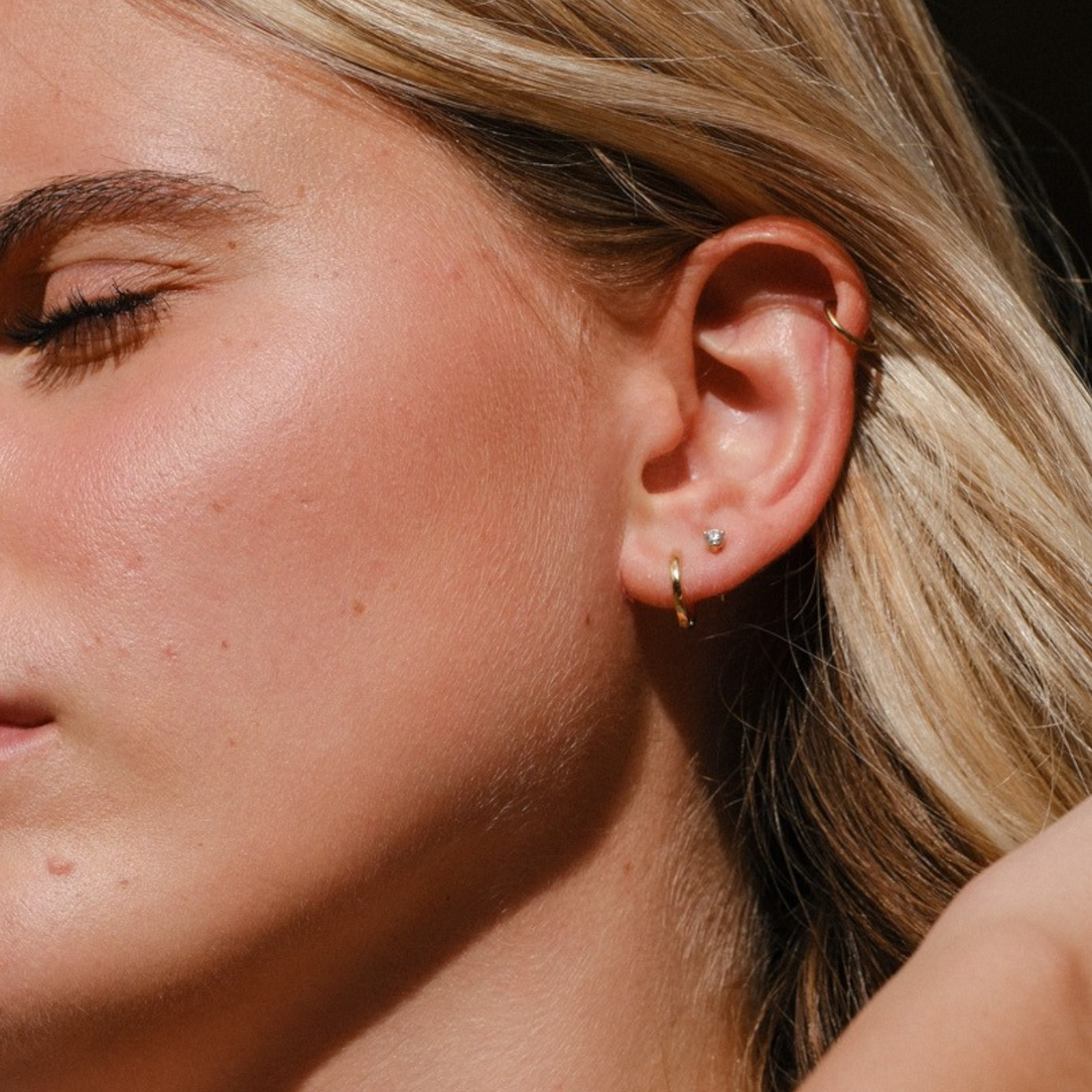 EC One single 18ct yellow gold huggie hoop earring worn by a woman with blonde hair in a close-up profile view.